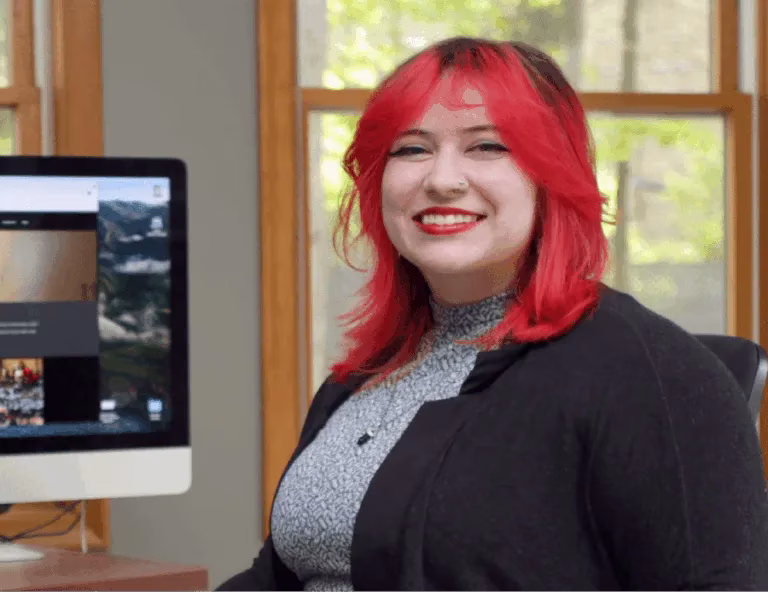 Smiling woman with bright red hair seated beside a desktop computer
